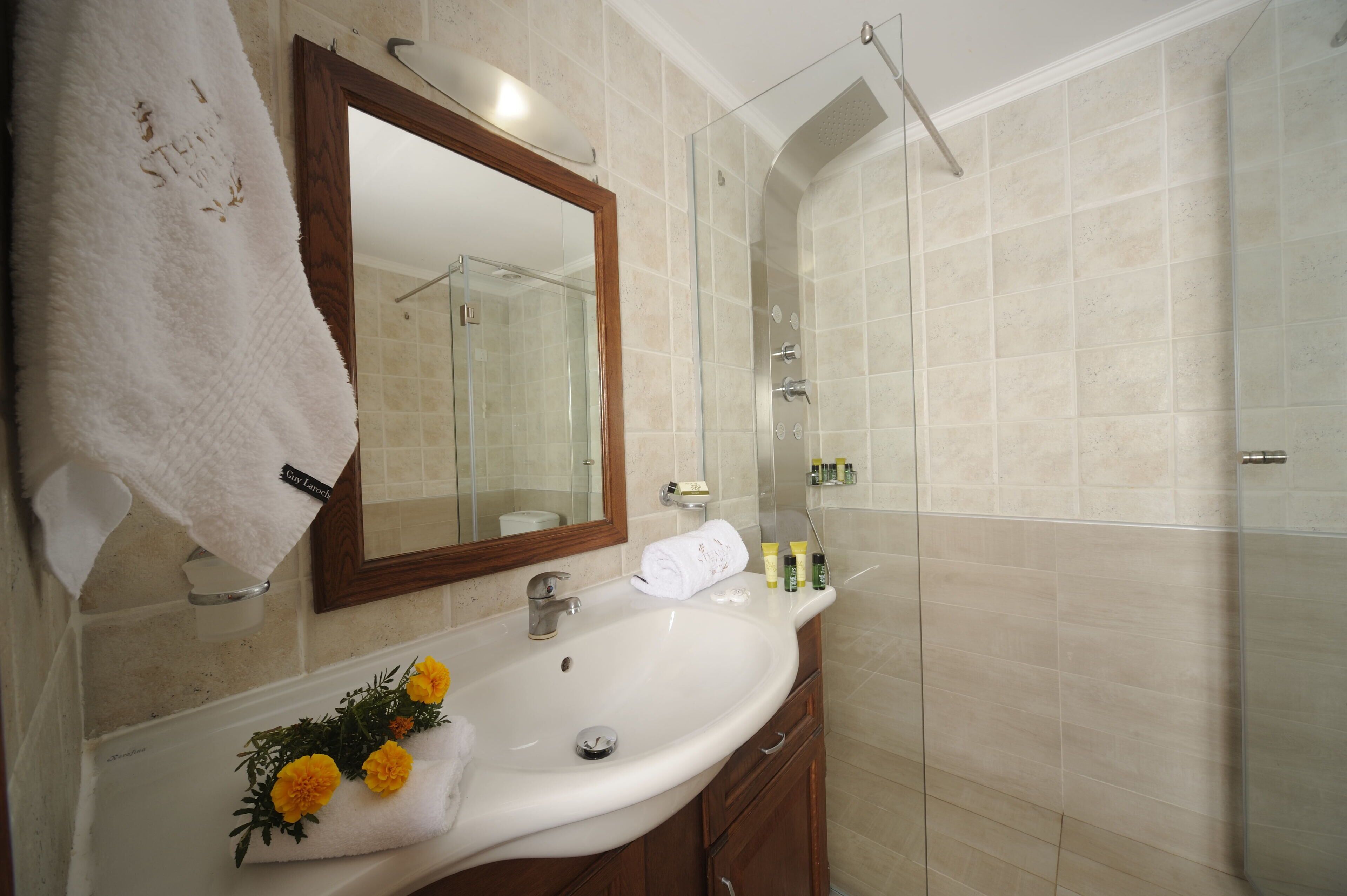 Un baño con lavamanos blanco, un espejo, cabina de ducha de vidrio y un perchero con una toalla blanca.