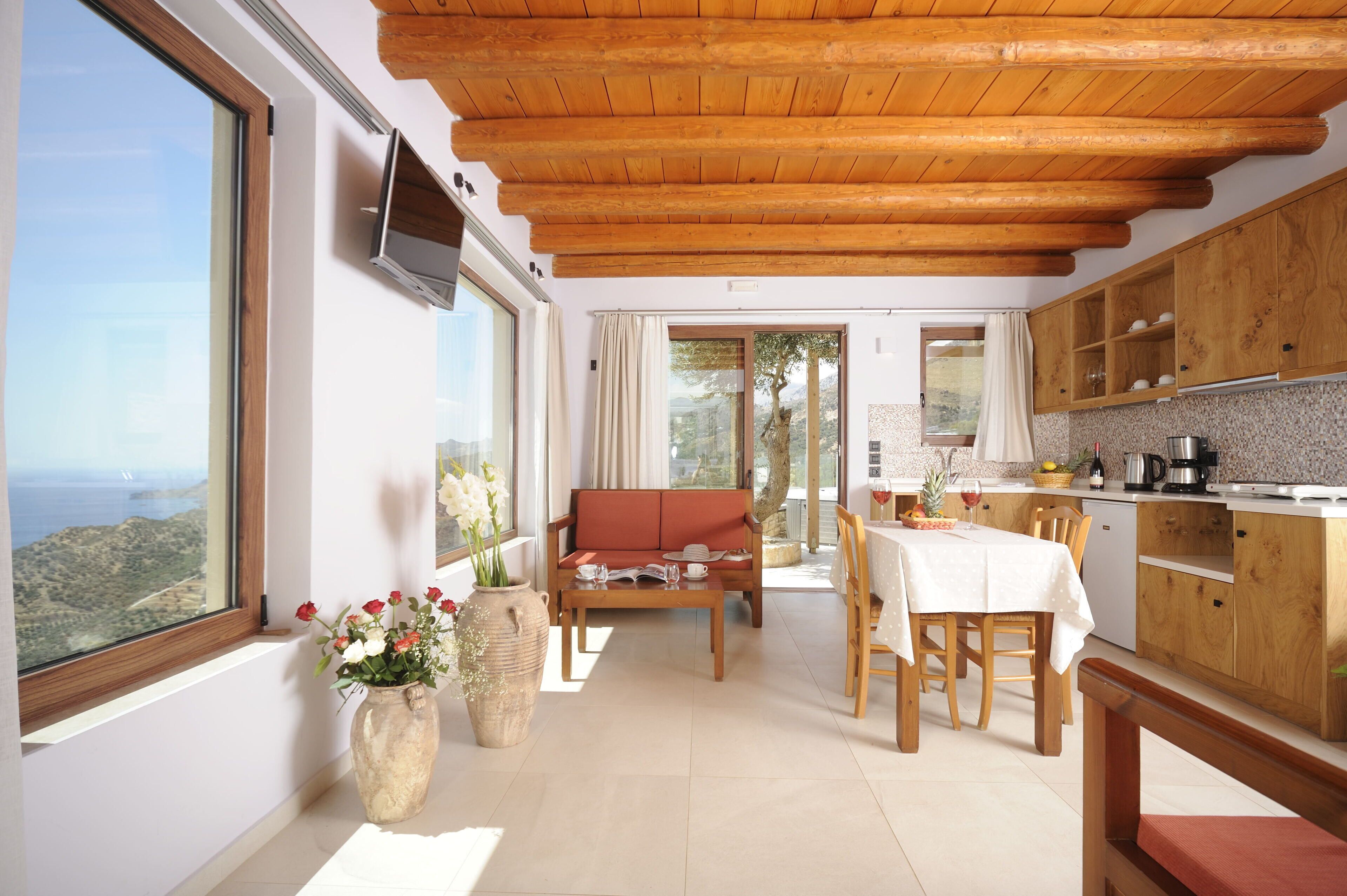A modern kitchen with wooden cabinets, a dining area with a table and chairs, a sofa, and a large window overlooking a scenic view.