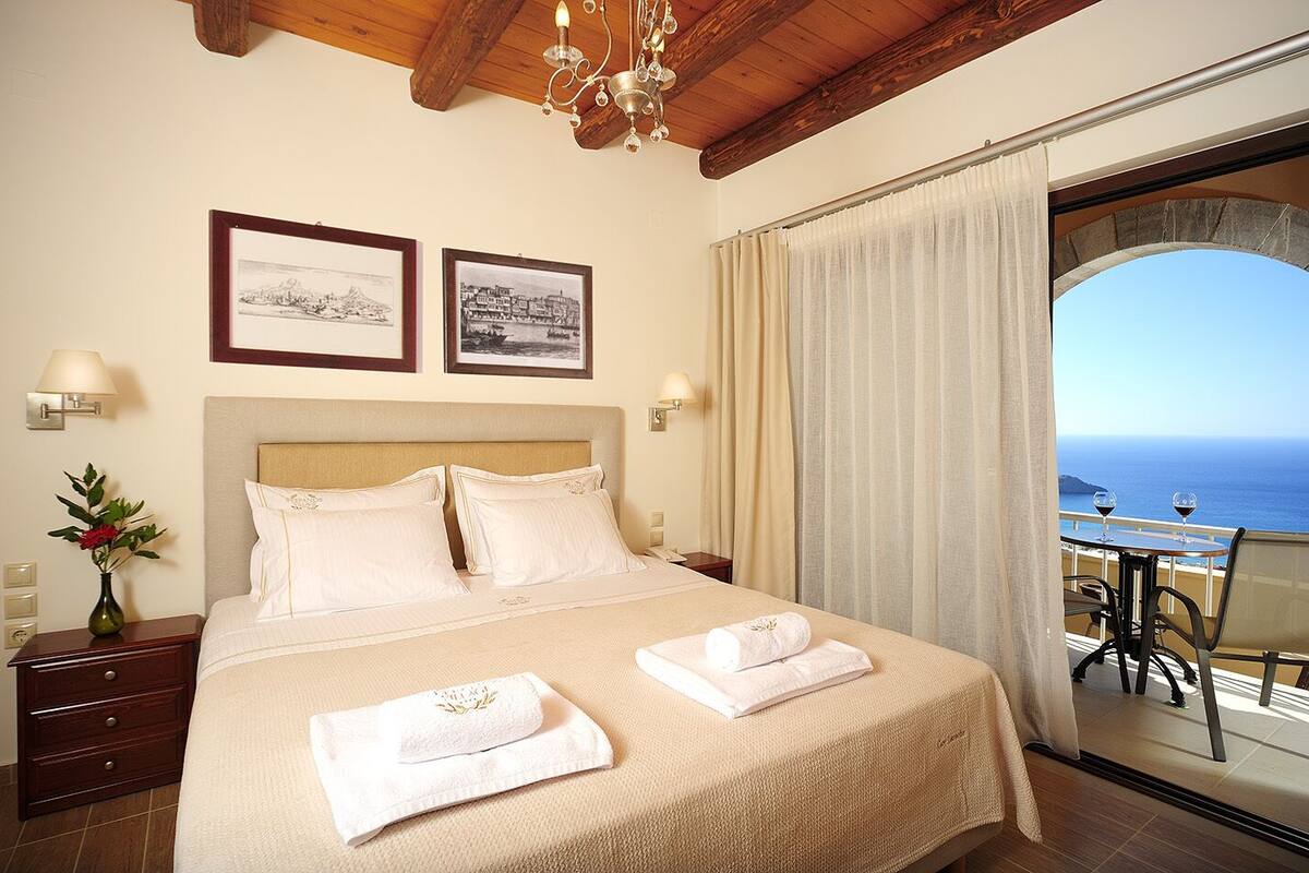 A hotel room with a bed, two framed pictures, a vase with flowers, a nightstand, and a balcony with a table and chairs.