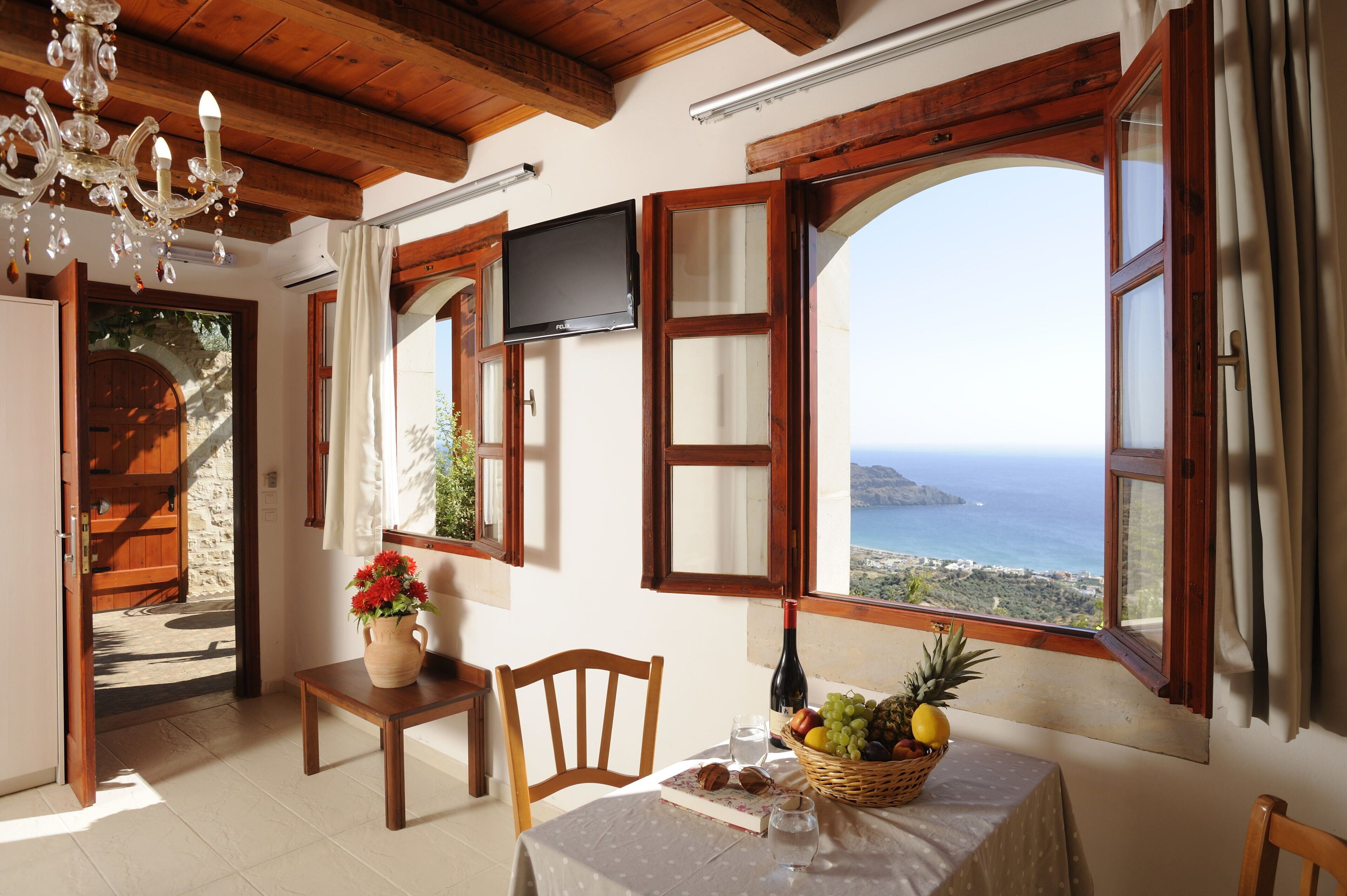 Une pièce avec un plafond en bois, un téléviseur fixé au mur, une table recouverte d’une nappe blanche et une vue sur la mer par de grandes fenêtres.