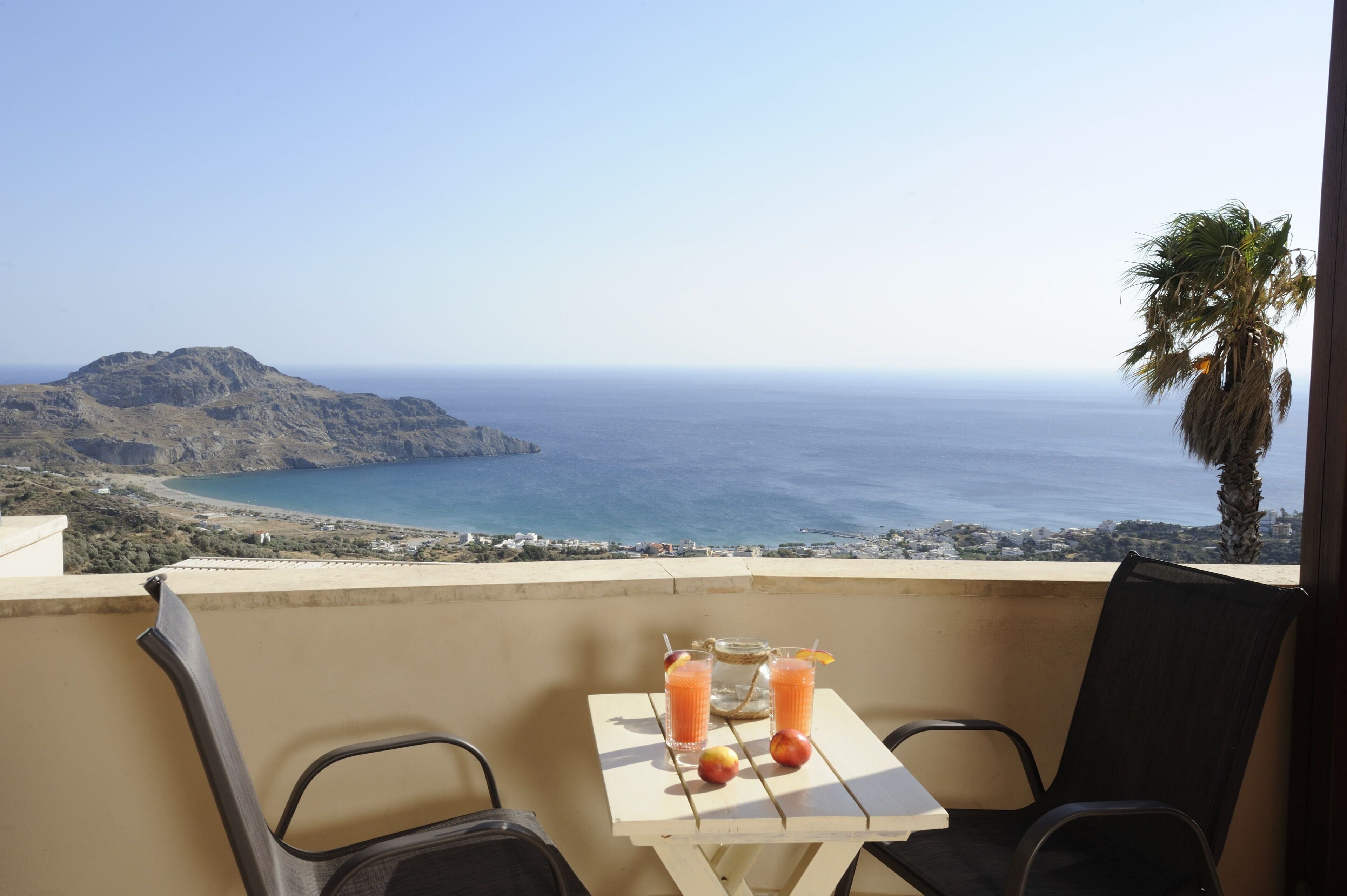 A balcony with a table set for two, overlooking a coastal landscape with a mountain and the sea.