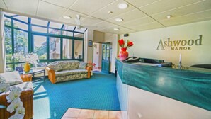 A reception area with a reception desk, a sofa, and a window providing a view of the outdoors. - Ashwood Manor Motor Lodge (Hamilton)