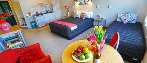 A compact hotel room with a bed, a small desk, a chair, and a dining table with fruit and a bottle.