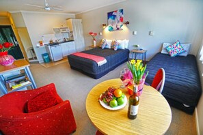 Ein kleines Hotelzimmer mit Bett, Schreibtisch, Stuhl und Esstisch mit Obst und einer Flasche.