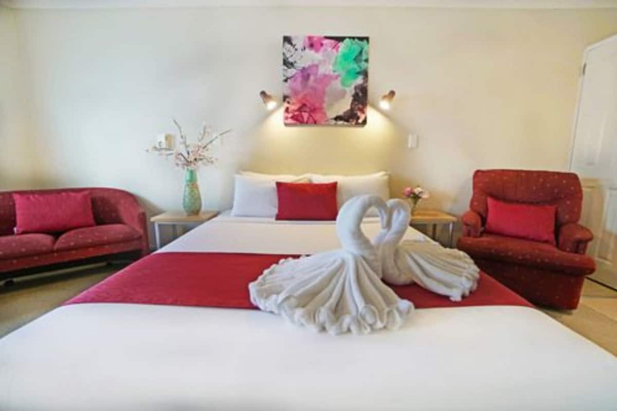 A hotel room with a bed, two red armchairs, a white swan-shaped towel, a vase with branches, and a colorful abstract painting on the wall.