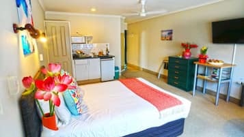 A bedroom with a bed, a desk, a chair, a dresser, a television, and a kitchen area.