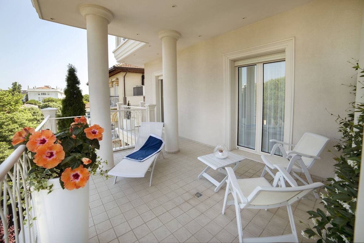 Un balcon avec des chaises blanches et une table, un pot de fleurs orné de fleurs oranges, et une vue sur les bâtiments environnants.