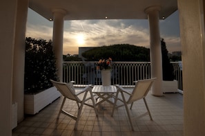 A balcony with a white table and chairs, a potted plant with flowers, and a view of the sunset over a hill. - Hotel Gala (Riccione)
