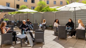 An outdoor seating area with wicker chairs and tables, umbrellas, and a building in the background. - a&o Wien Hauptbahnhof (Vienna)