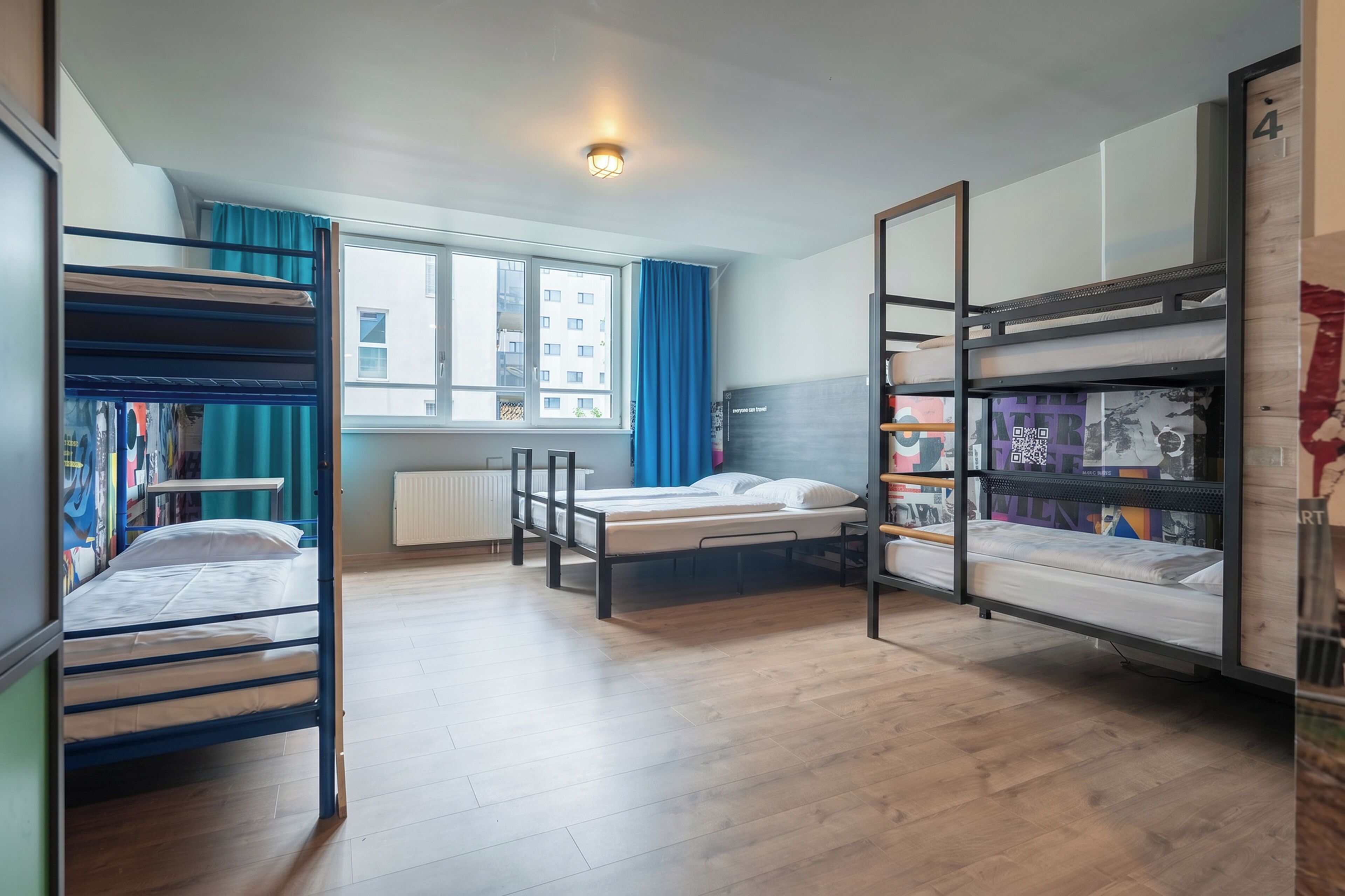 A dormitory room with bunk beds, a window with curtains, a radiator, and a bookshelf.