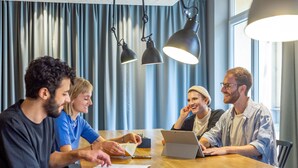 A group of people sitting around a wooden table in a room with blue curtains and hanging pendant lights. - a&o Wien Hauptbahnhof (Vienna)