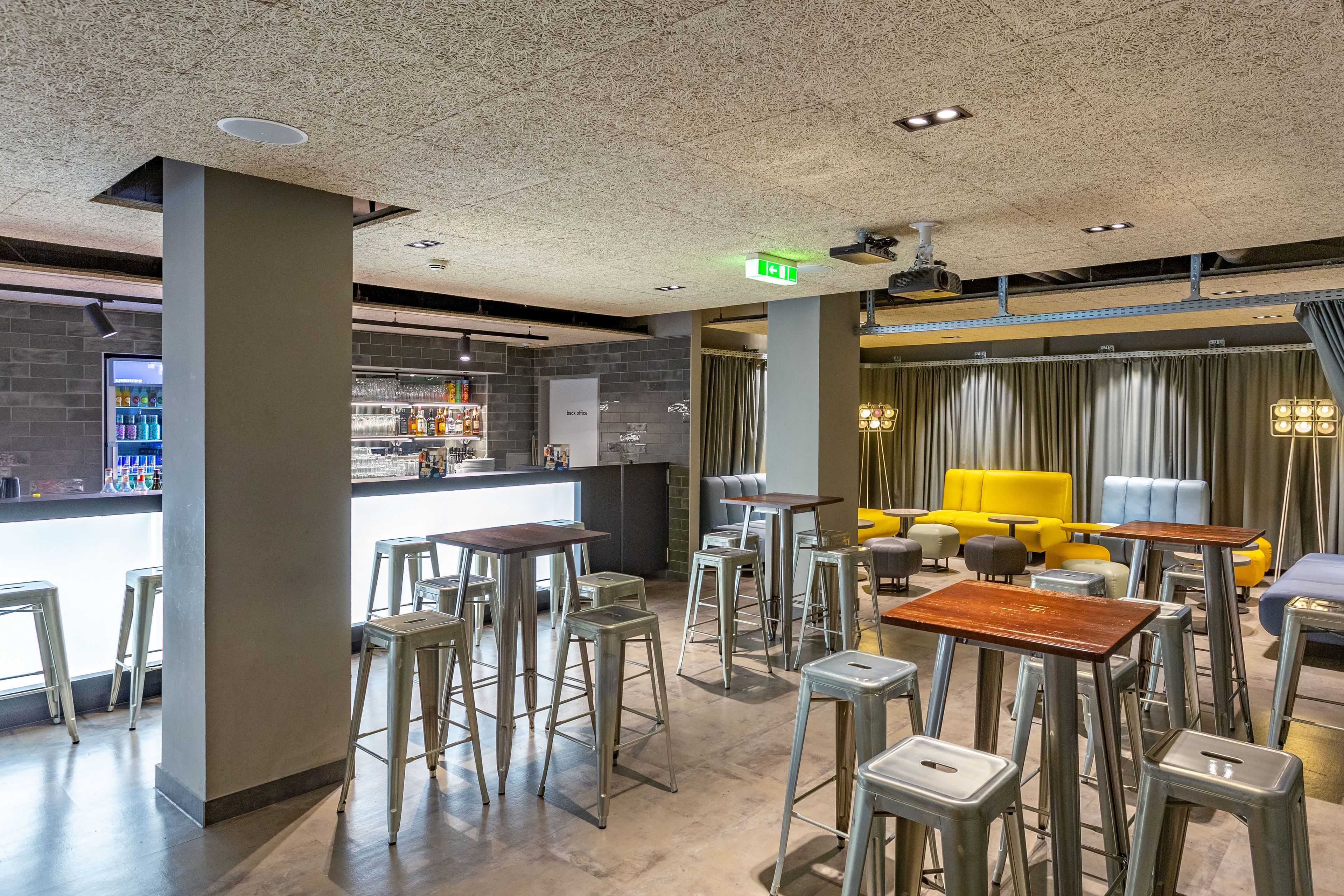 A modern bar area with stools and tables, a counter with beverages, and a seating area with yellow and blue couches.