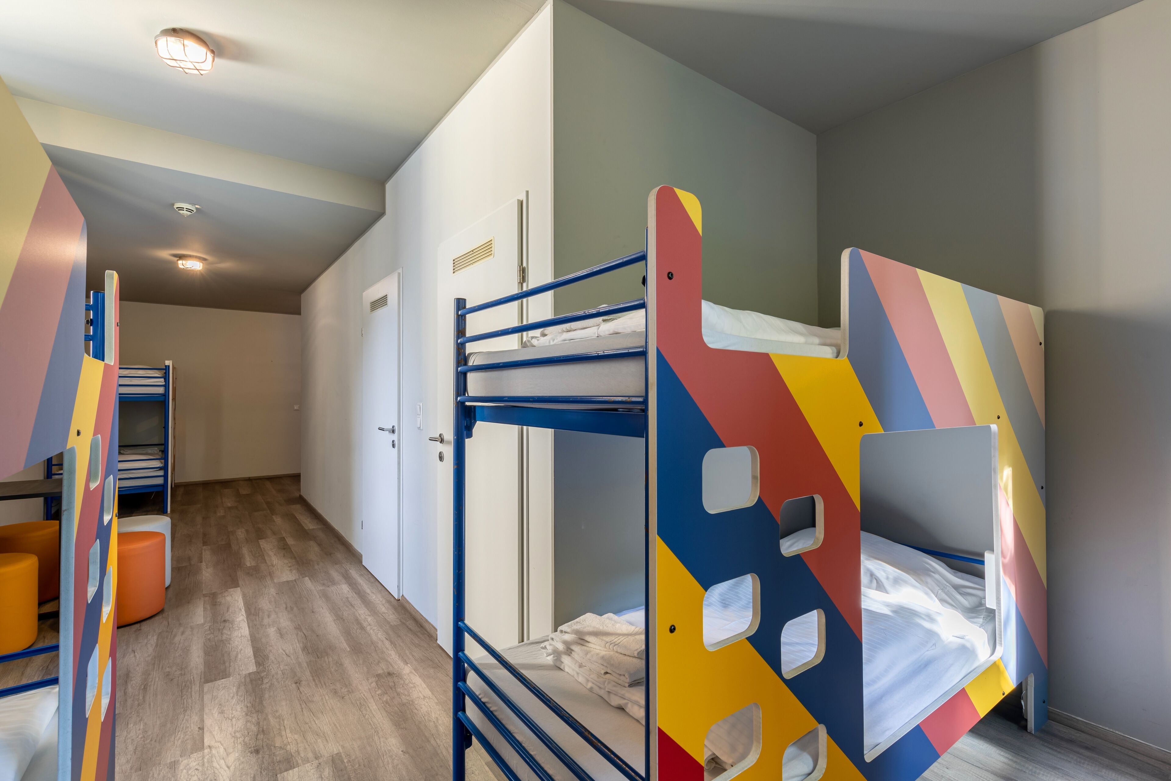 A bunk bed room with a colorful headboard, a wardrobe, and a small seating area with orange cushions.
