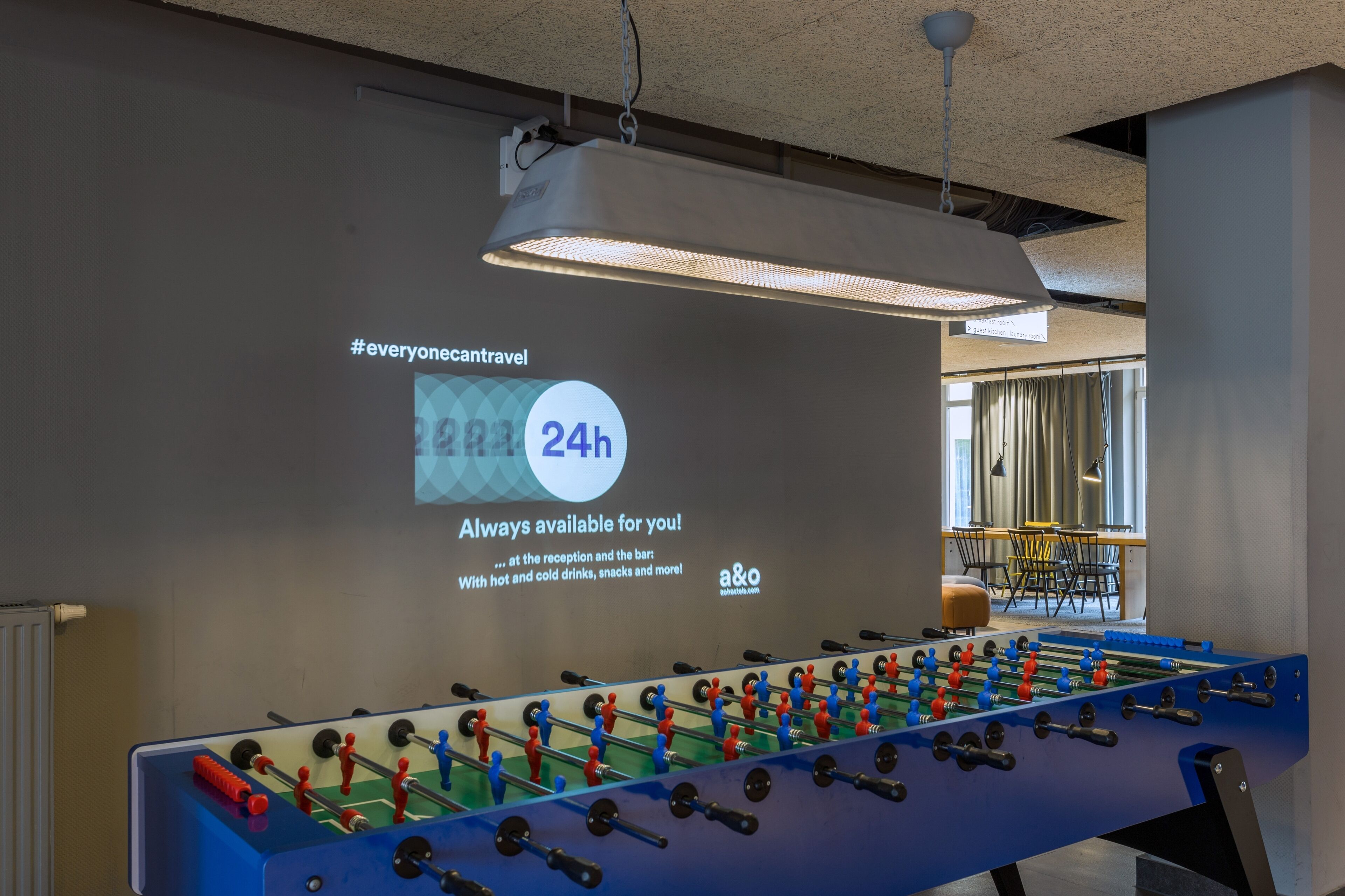 A foosball table in a room with a projection screen displaying a message about 24-hour availability.