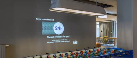 A foosball table in a room with a projection screen displaying a message about 24-hour availability.