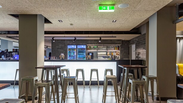 A modern bar area with stools and a counter, featuring a brick wall and a visible exit sign. - a&o Wien Hauptbahnhof (Vienna)