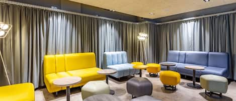 A modern lounge area with sofas and ottomans in yellow, blue, and gray, arranged around small round tables.