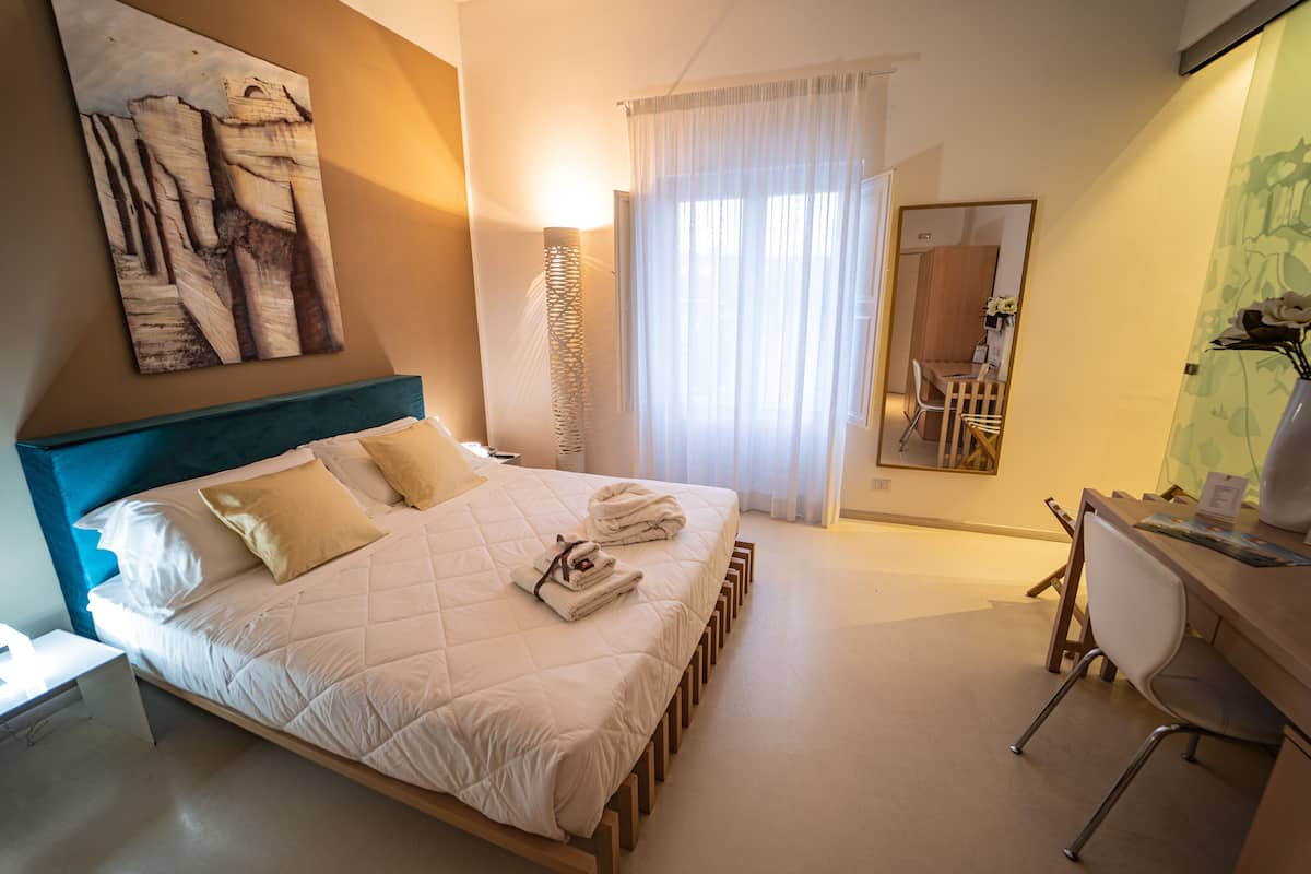 A hotel room with a large bed, a desk, a chair, a mirror, and a painting on the wall.