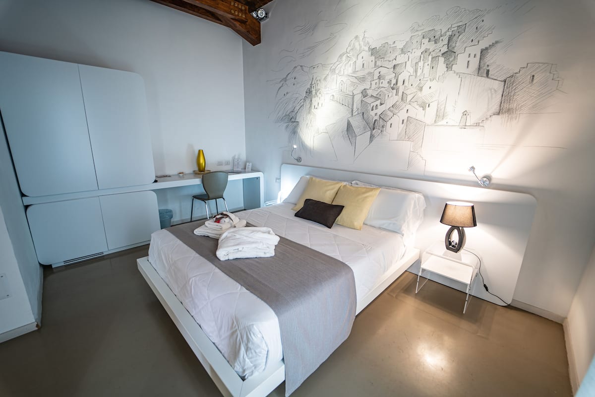 A modern hotel room with a large bed, a desk, and a chair. There is a wall mural and a bedside lamp.