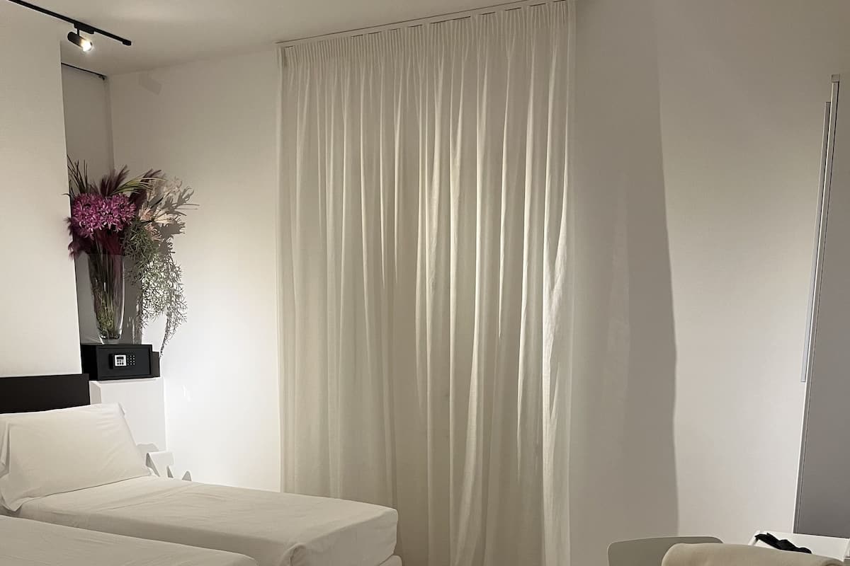 A modern hotel room with two white beds, a desk with a chair, a vase with flowers, and a large window with white curtains.