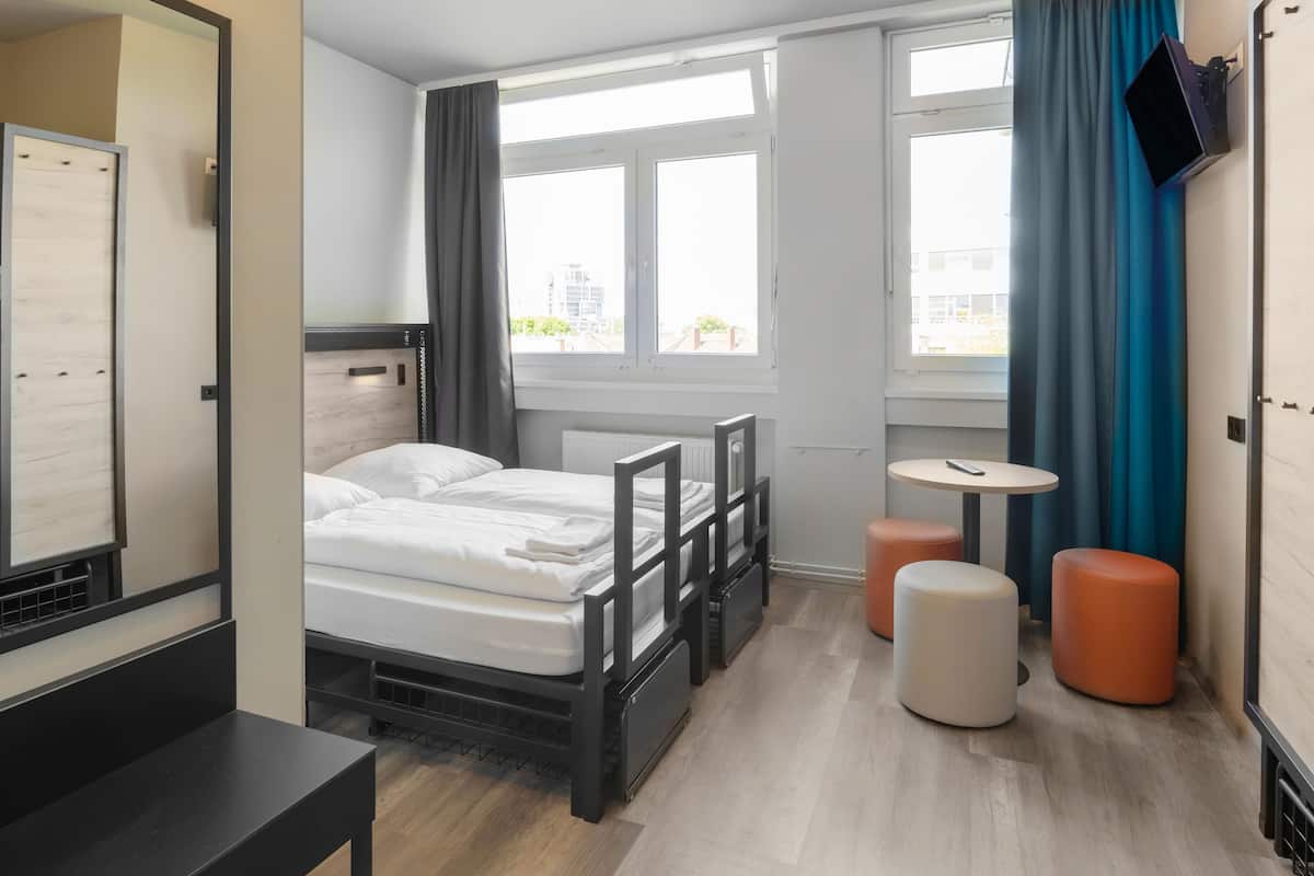 Une chambre d’hôtel moderne équipée d’un lit, d’une petite table, de deux poufs, d’un téléviseur fixé au mur et de grandes fenêtres avec des rideaux.