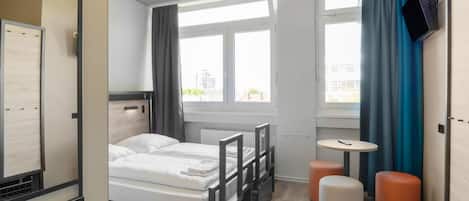 Una moderna camera d'albergo con un letto, un tavolino, due pouf, una TV a parete e ampie finestre con tende.