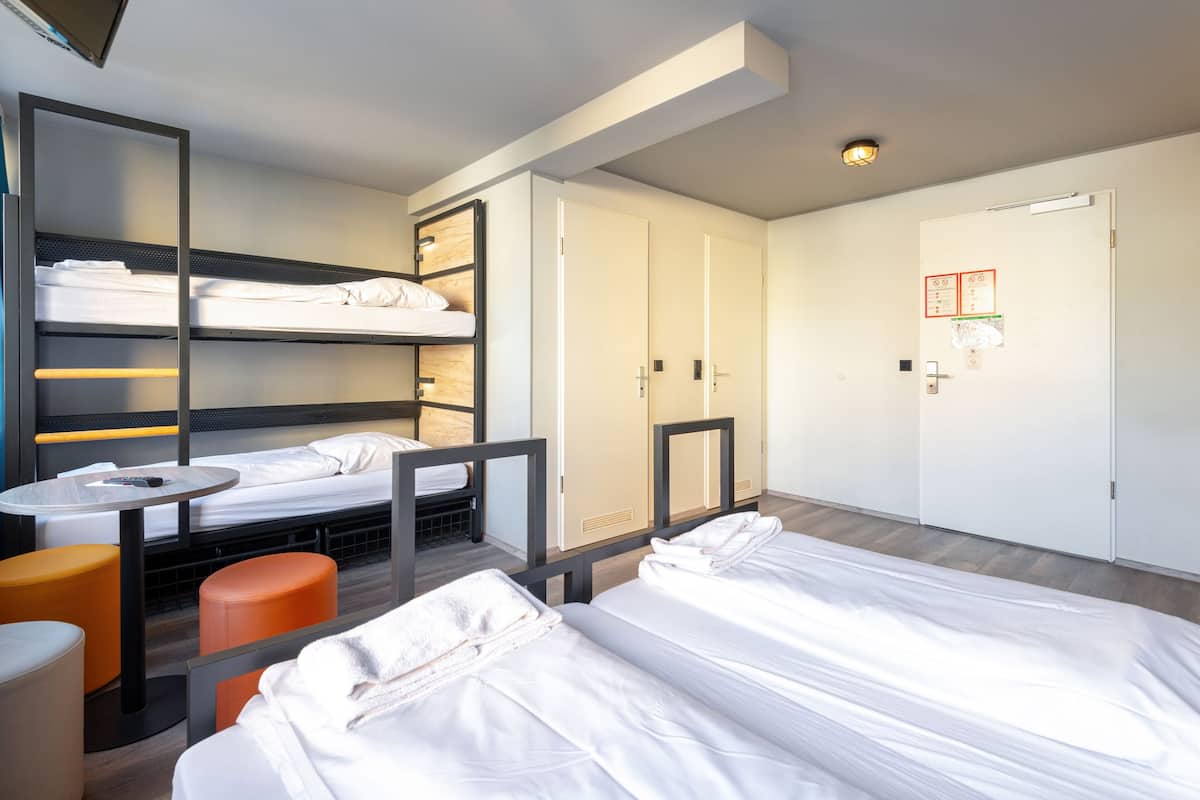 Une chambre d’hôtel avec des lits superposés, une petite table, un pouf orange et une porte blanche sur laquelle est affiché un avis.