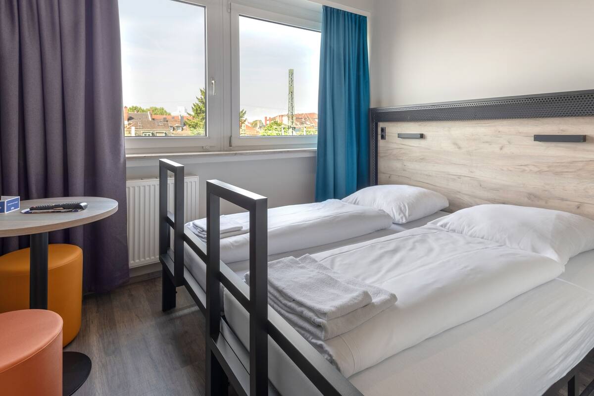 Une chambre d’hôtel avec deux lits, une petite table, une chaise et une fenêtre avec des rideaux.