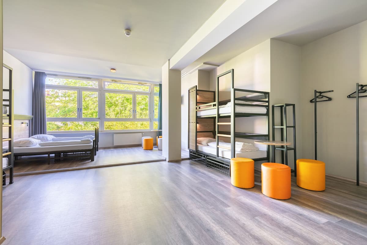 Une chambre d’hôtel moderne avec une grande fenêtre, des lits superposés et des poufs orange.