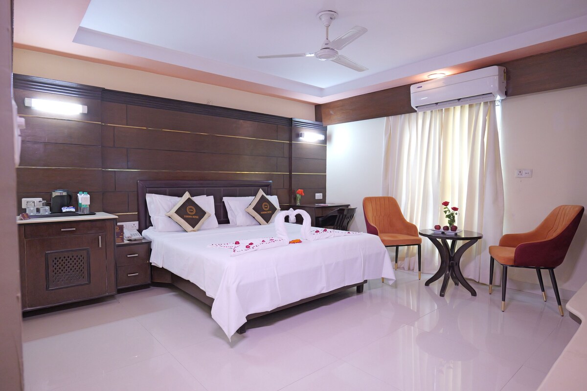 A hotel room with a bed, bedside tables, a desk, two chairs, a ceiling fan, and an air conditioning unit.