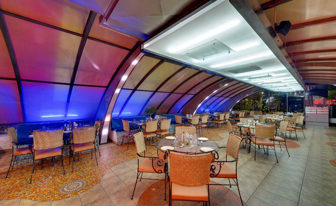 A spacious dining area with multiple tables and chairs, a curved ceiling, and a colorful floor pattern.