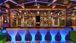 A bar area with wooden beams, hanging lights, and a brick wall. There are illuminated stools and a bar counter with bottles. - Hotel Combermere (Shimla)