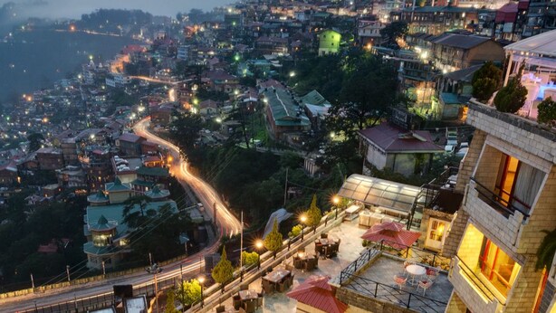 A rooftop terrace with outdoor seating, a view of a cityscape, and a prominent tower structure. - Hotel Combermere (Shimla)