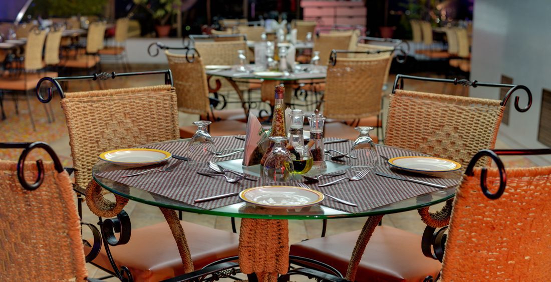 A round glass table with a woven chair backrest, set with plates, cutlery, and condiment bottles.