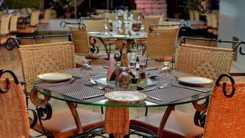 A round glass table with a woven chair backrest, set with plates, cutlery, and condiment bottles.