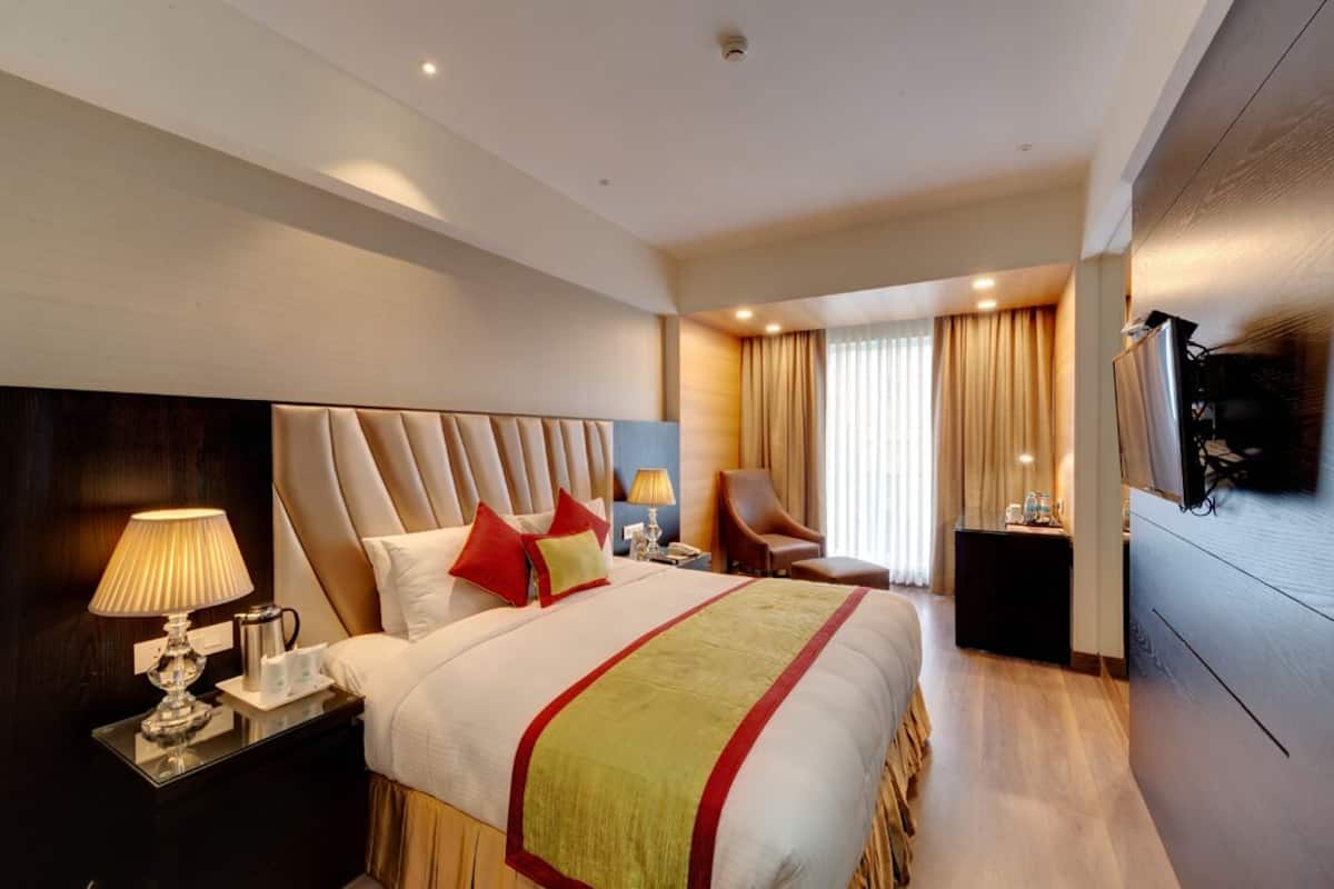 A hotel room with a large bed, a bedside table with a lamp, a chair, a TV mounted on the wall, and a window with curtains.