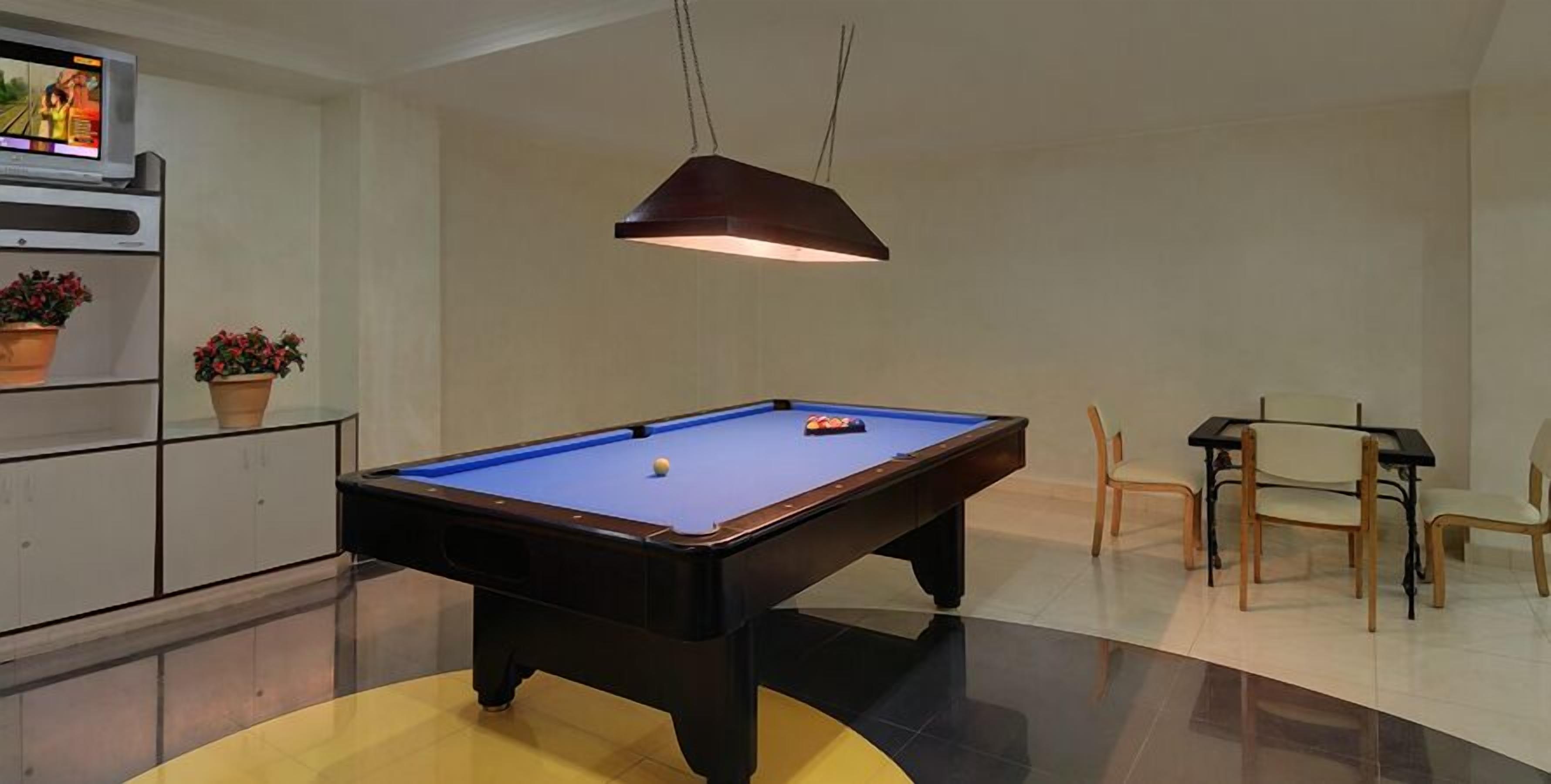 A pool table with a single ball, a television displaying a sports game, and a dining area with chairs and a table.