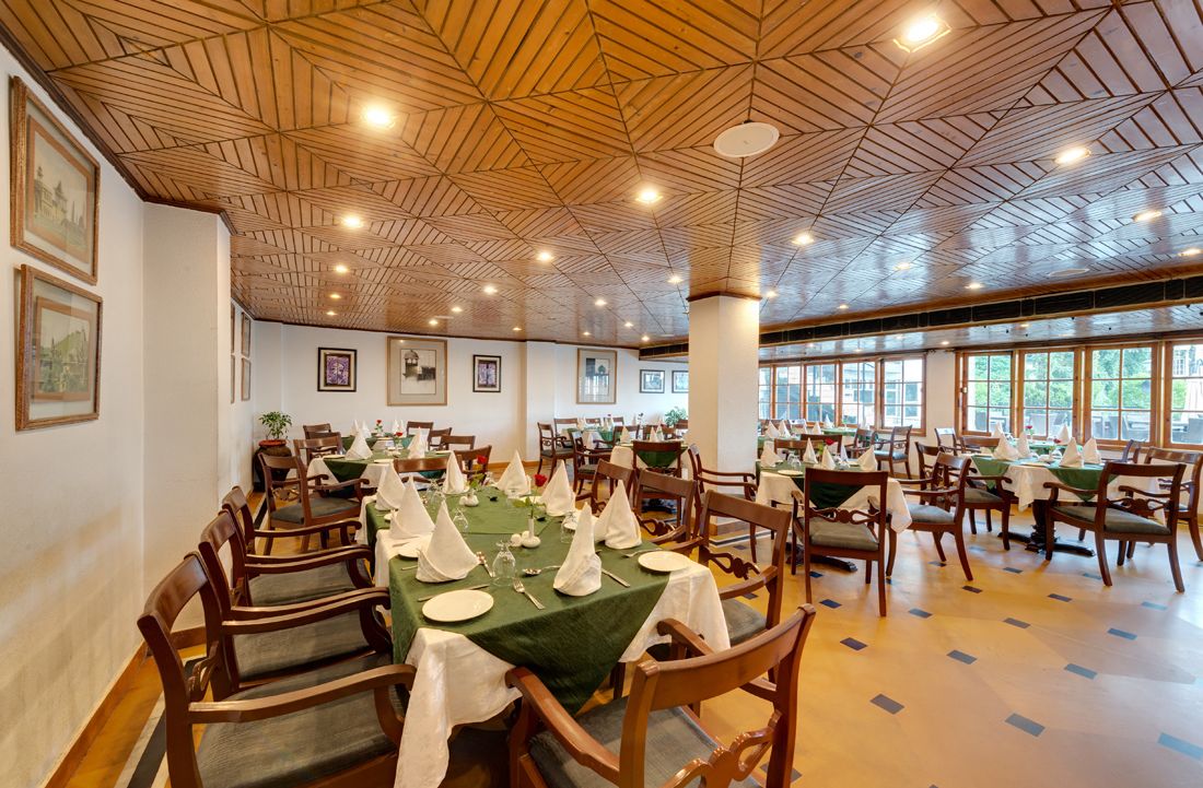 A dining area with wooden ceiling, tables set with white tablecloths and napkins, and chairs with green cushions.