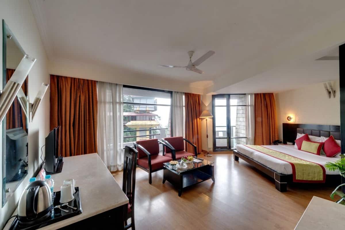 A hotel room with a bed, a desk with a kettle, a chair, a coffee table, and a large window with curtains.