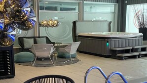 Un área de piscina interior moderna con sillas negras, una mesa de vidrio con una vela y una bañera de hidromasaje al fondo.