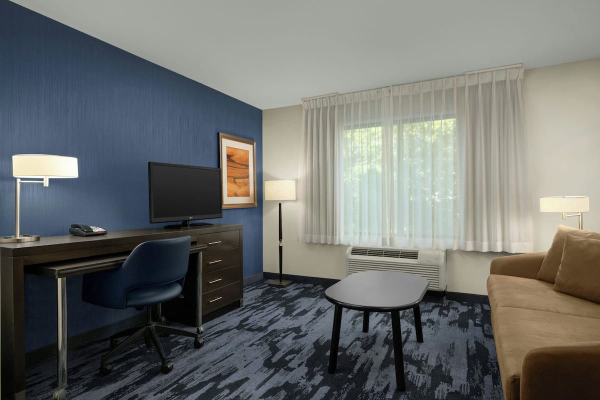 A hotel room with a desk, chair, television, two lamps, a sofa, and a large window with curtains.