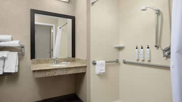 A modern bathroom with a granite countertop sink, a mirror, a shower with a handheld showerhead, and a grab bar. There are towels and toiletries on the counter.