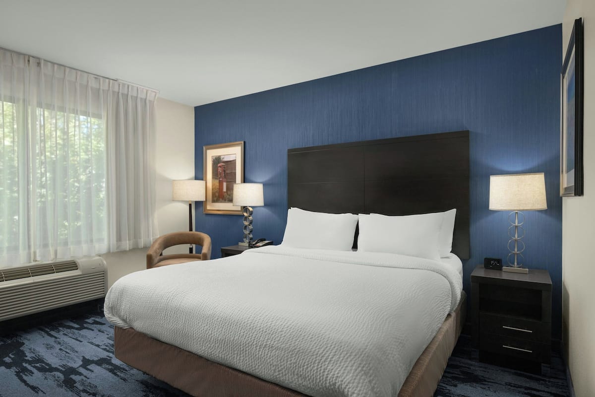 A hotel room with a large bed, a nightstand with a lamp, a chair, and a window with curtains.