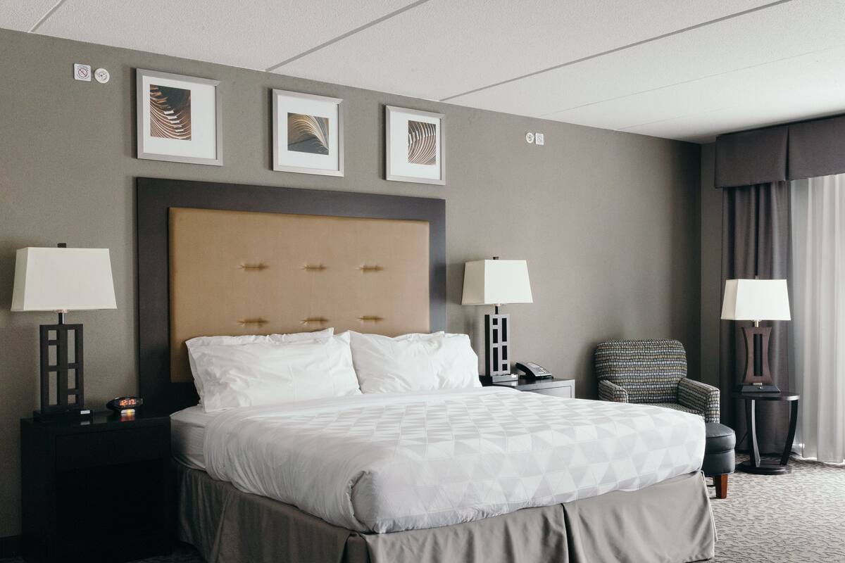 Une chambre d’hôtel avec un grand lit, deux tables de chevet avec des lampes, une chaise et des œuvres d’art encadrées au mur.