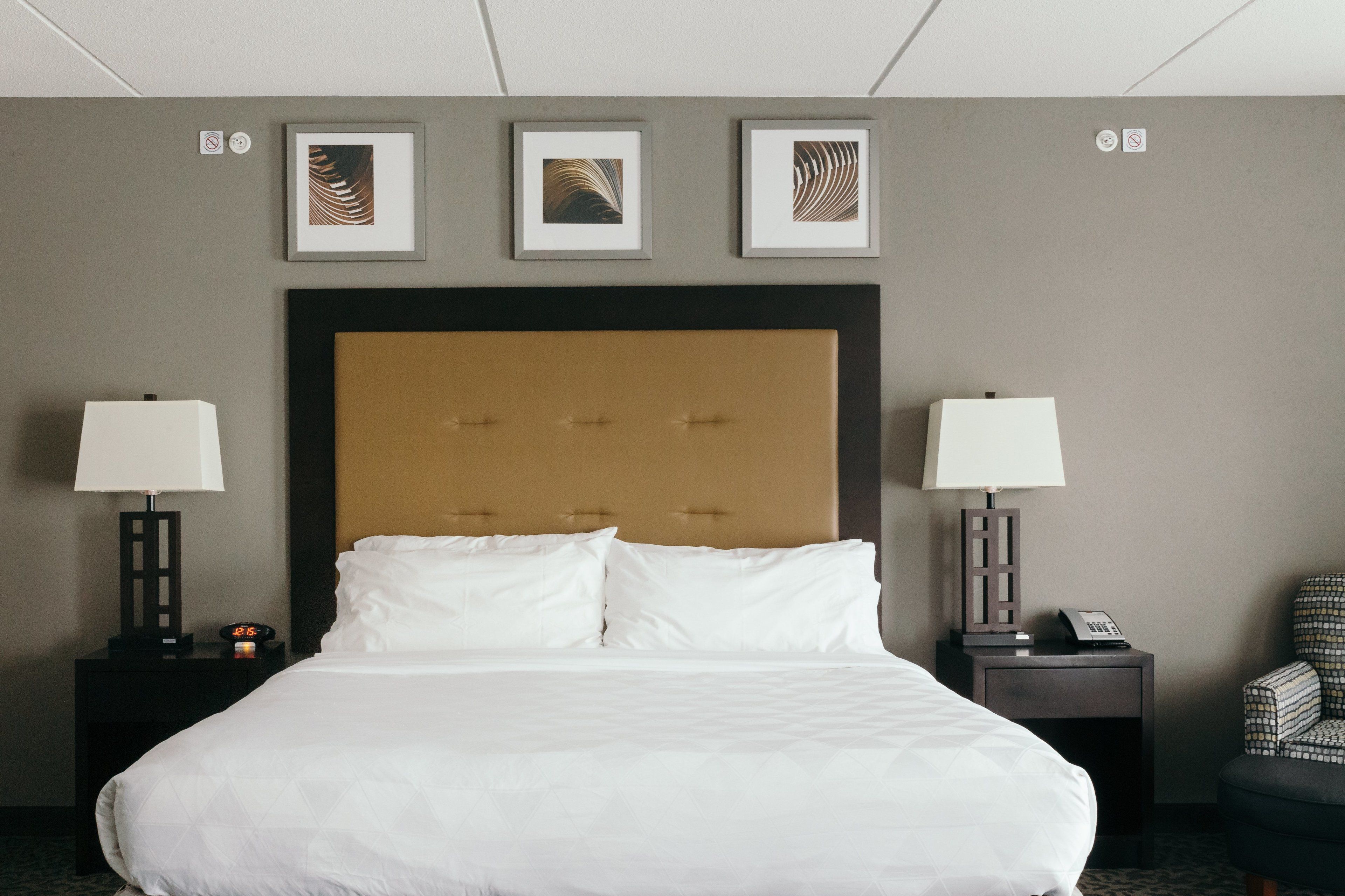A hotel room with a large bed, two bedside tables with lamps, a chair, and three framed artworks on the wall.