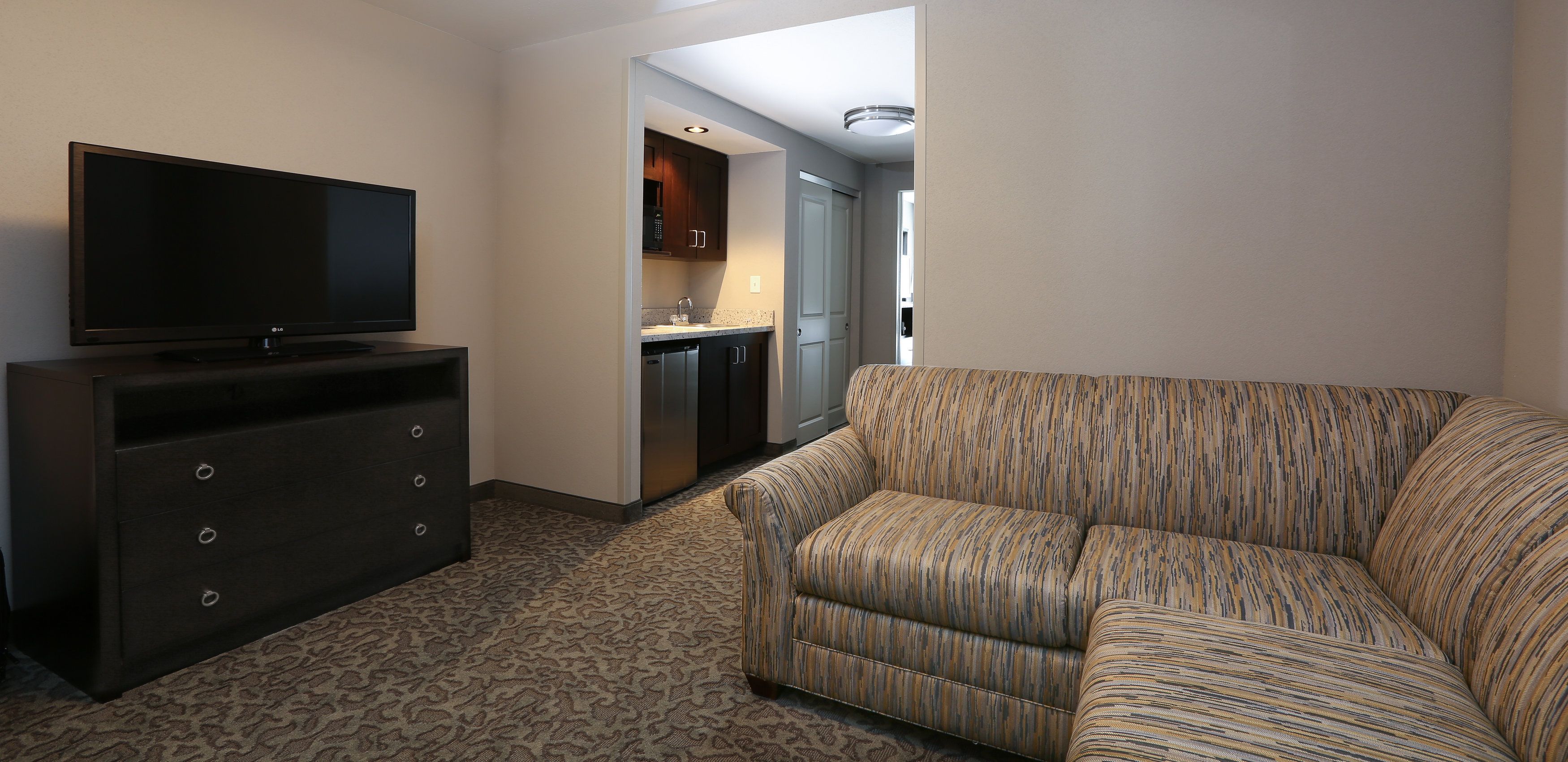 Ett modernt hotellrum med en platt-TV placerad på en mörk träbyrå, en mönstrad soffa och ett litet kök i bakgrunden.