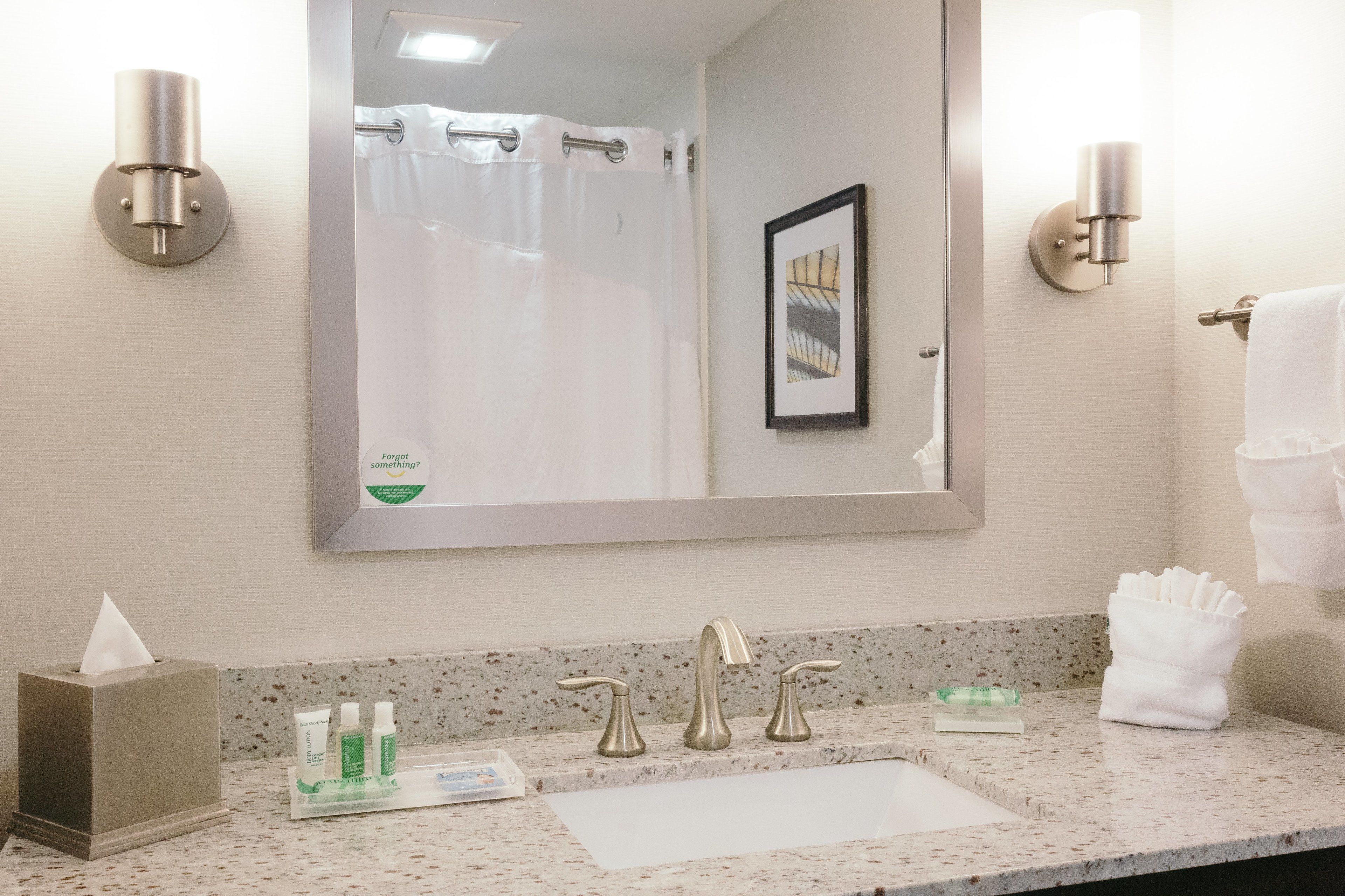 A bathroom with a large mirror, a sink, two wall-mounted lights, a towel rack, and a framed picture on the wall.
