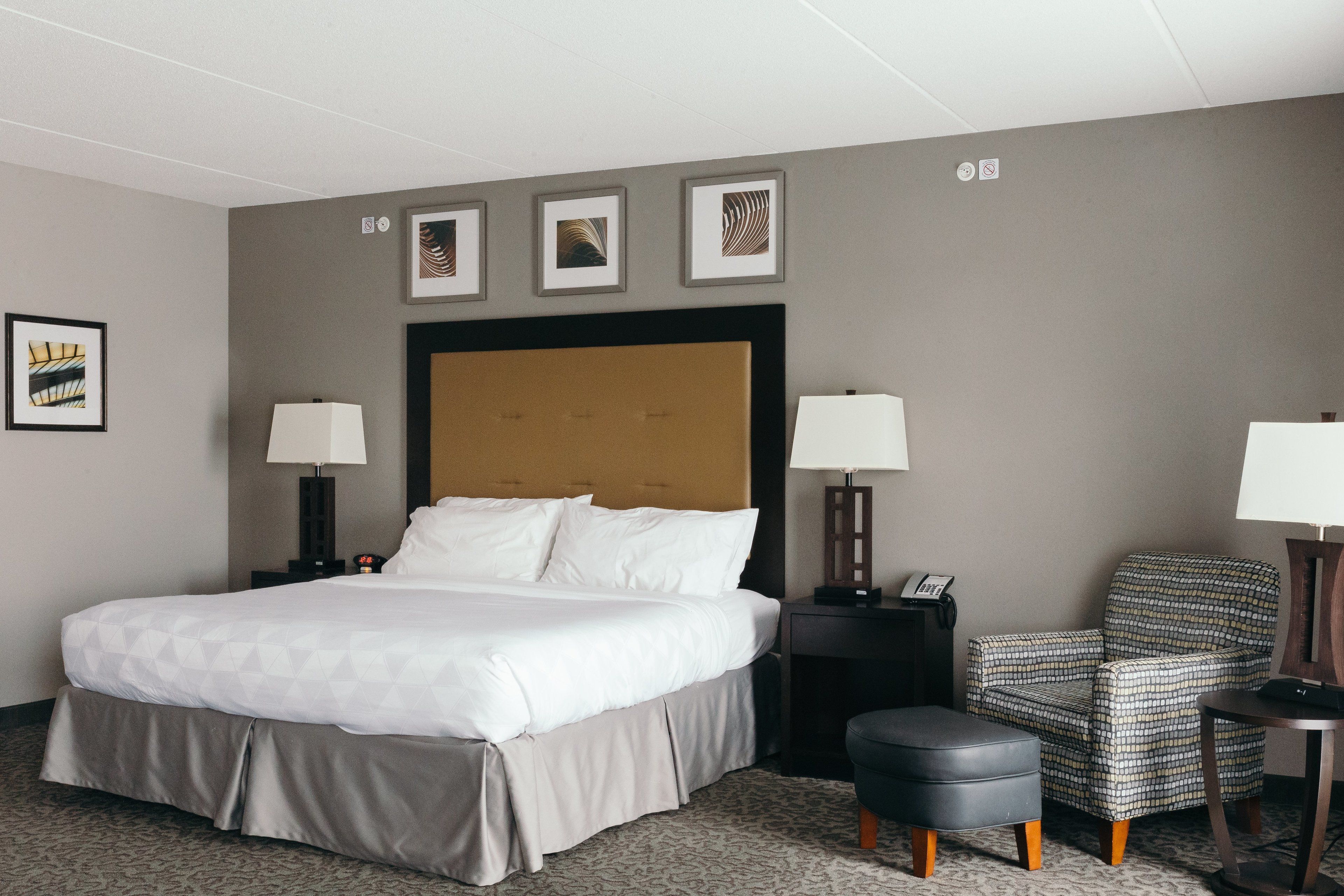 A hotel room with a large bed, two bedside tables with lamps, a chair, a small ottoman, and framed artwork on the wall.