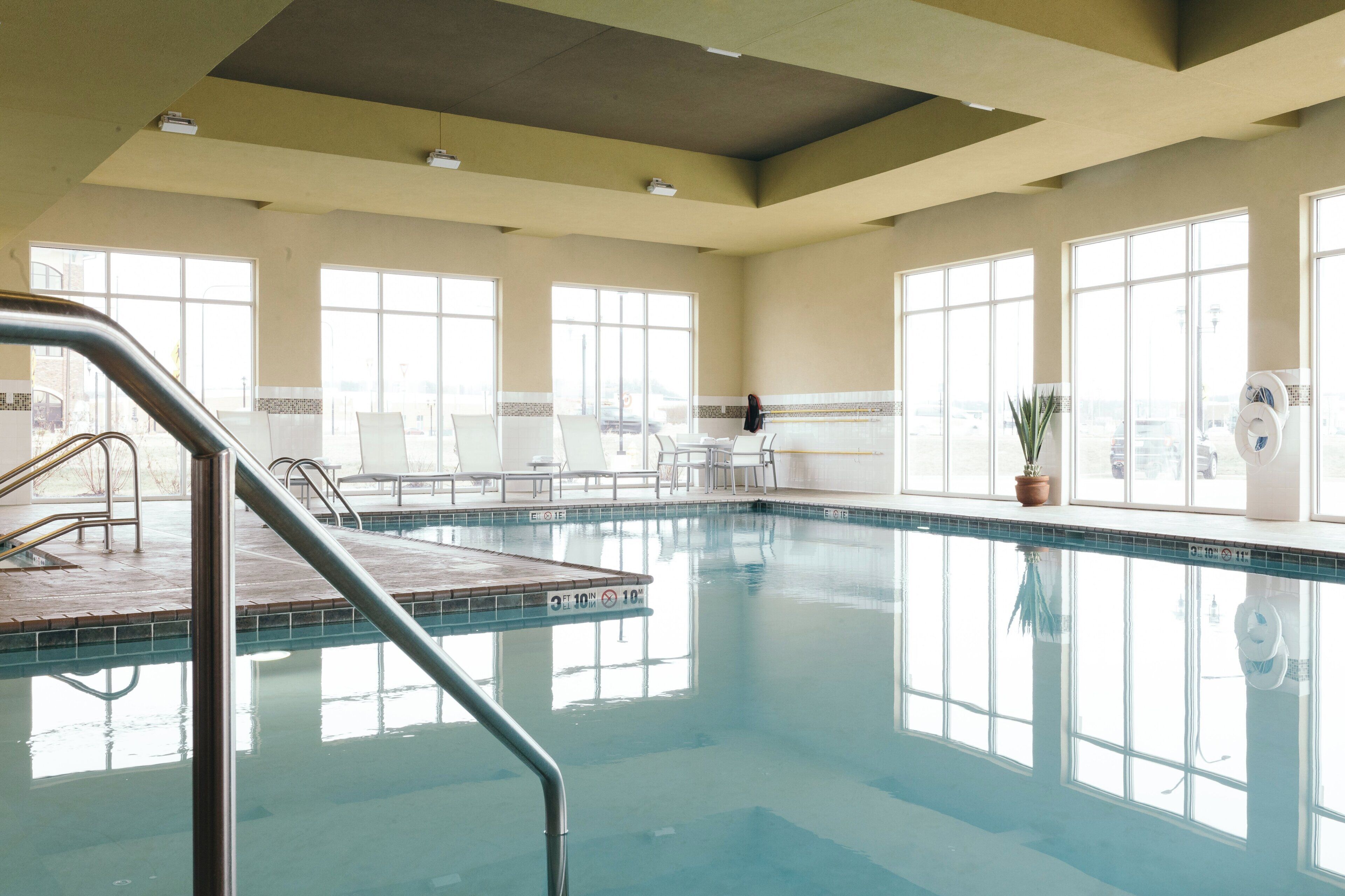A large indoor swimming pool with clear blue water, surrounded by windows and a poolside area with chairs and a small table.