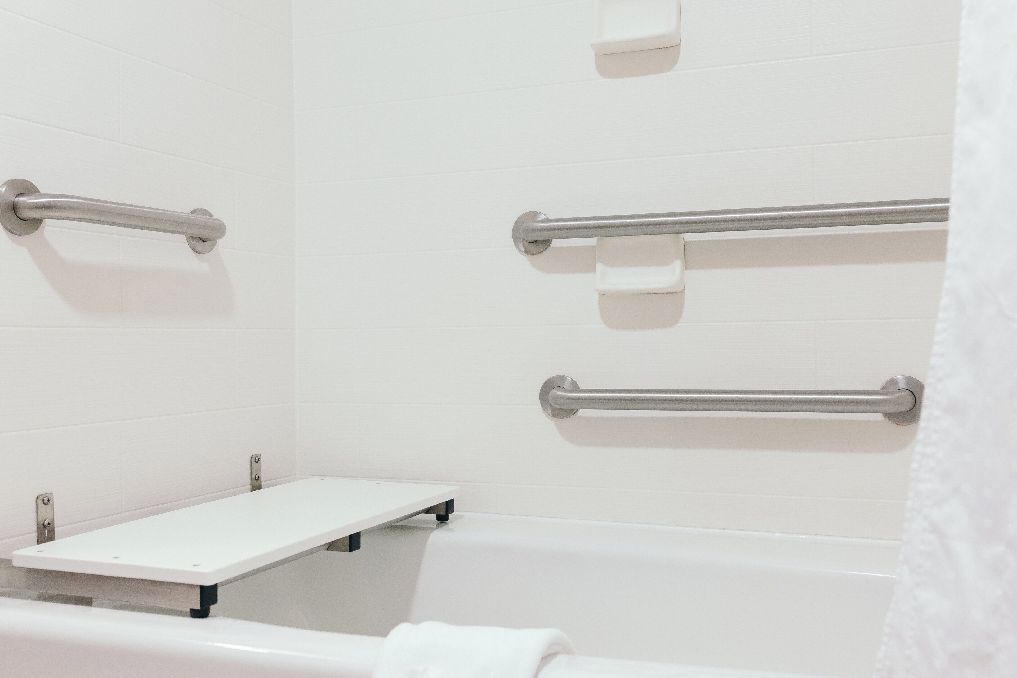 A bathroom with a white bathtub, a white sink, and a white shower area. There are three grab bars installed on the walls.